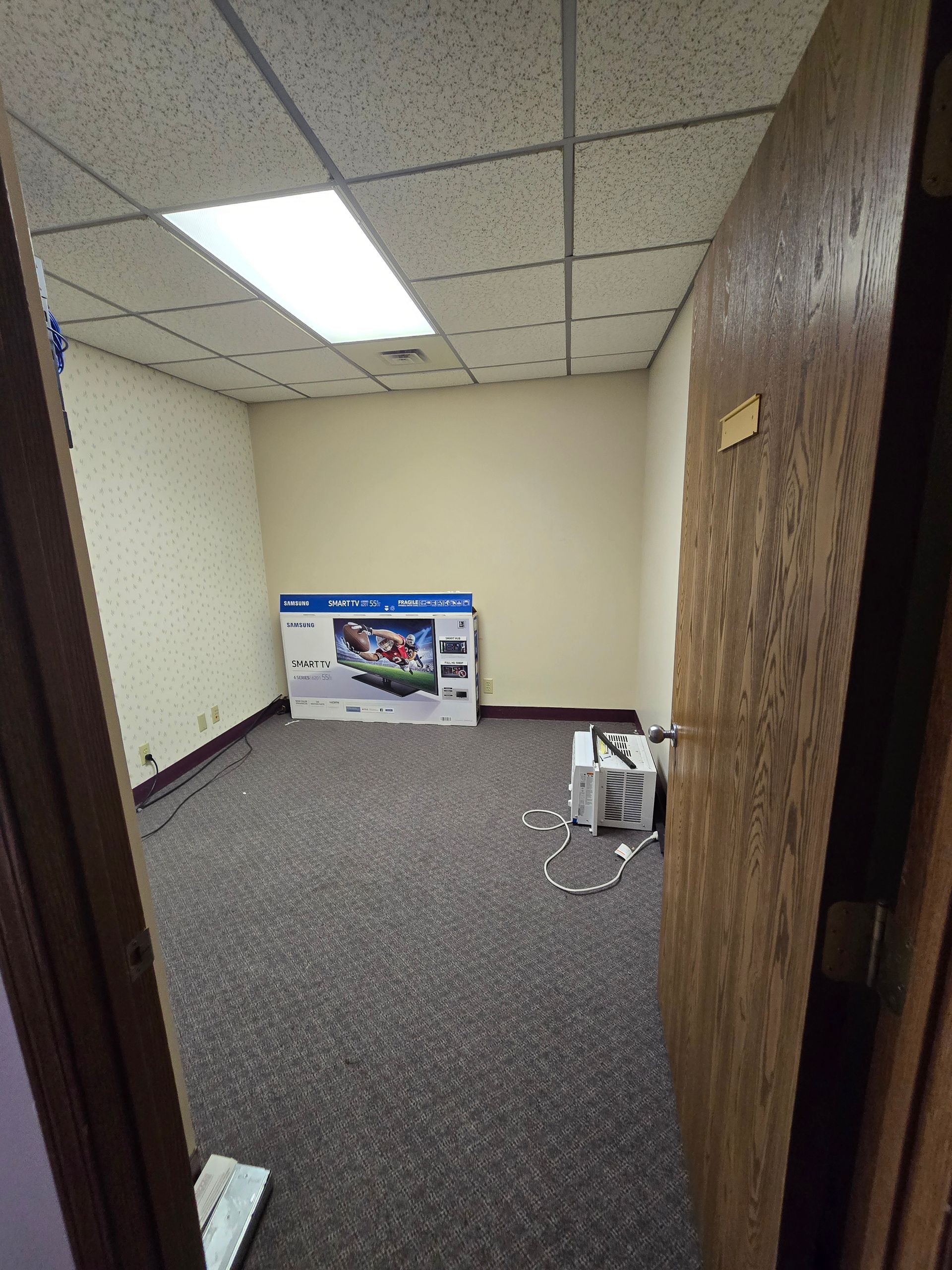 Empty office hallway with gray carpet, beige walls, ceiling tiles, and a poster leaning against the wall.