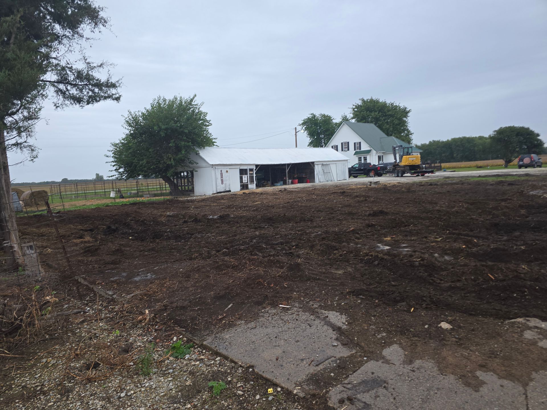 Cleared dirt lot with a white house, trees, and a yellow excavator under a cloudy sky