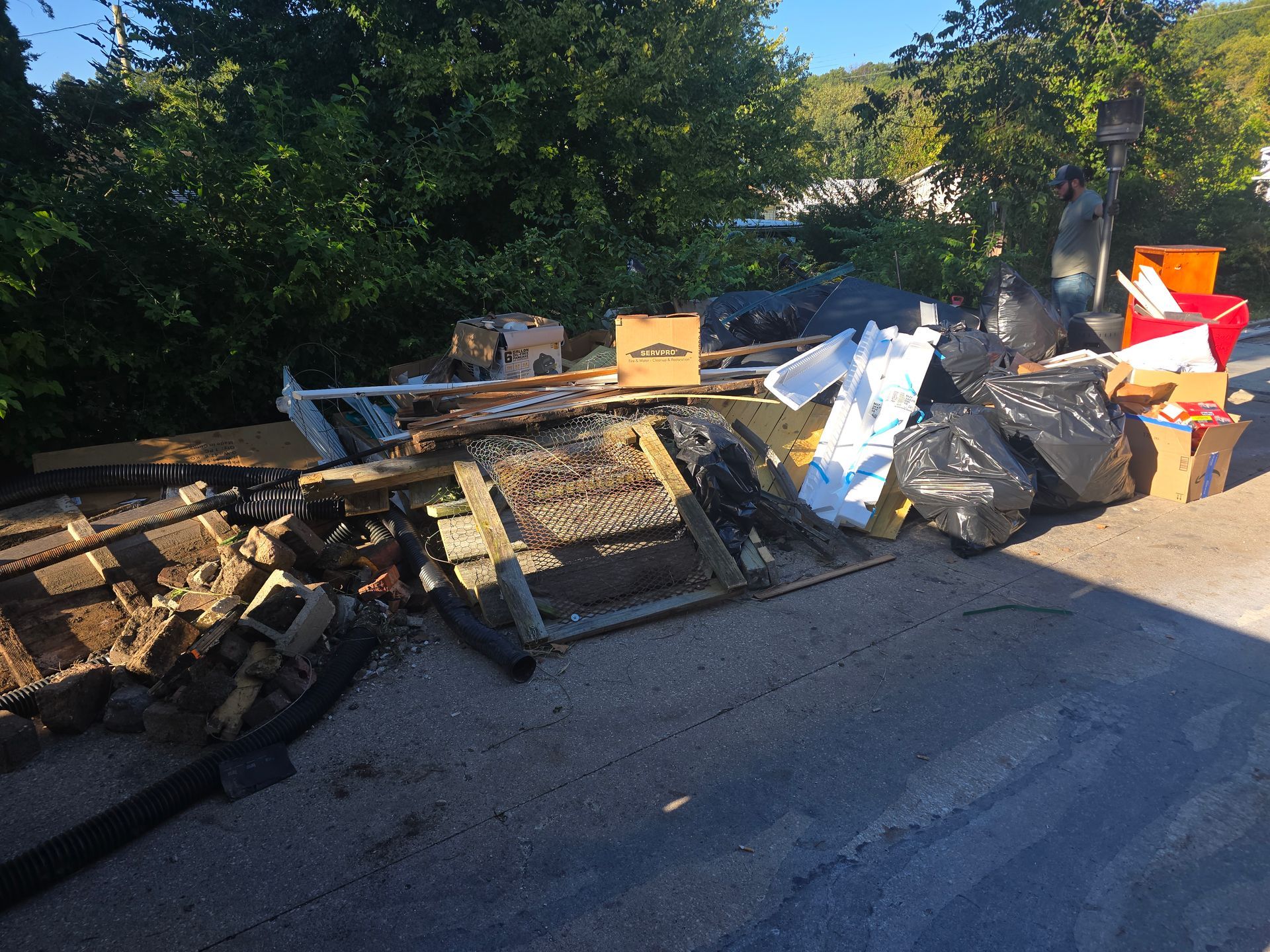 Piles of debris and flattened metal frames piled along a roadside beside trees and bushes.