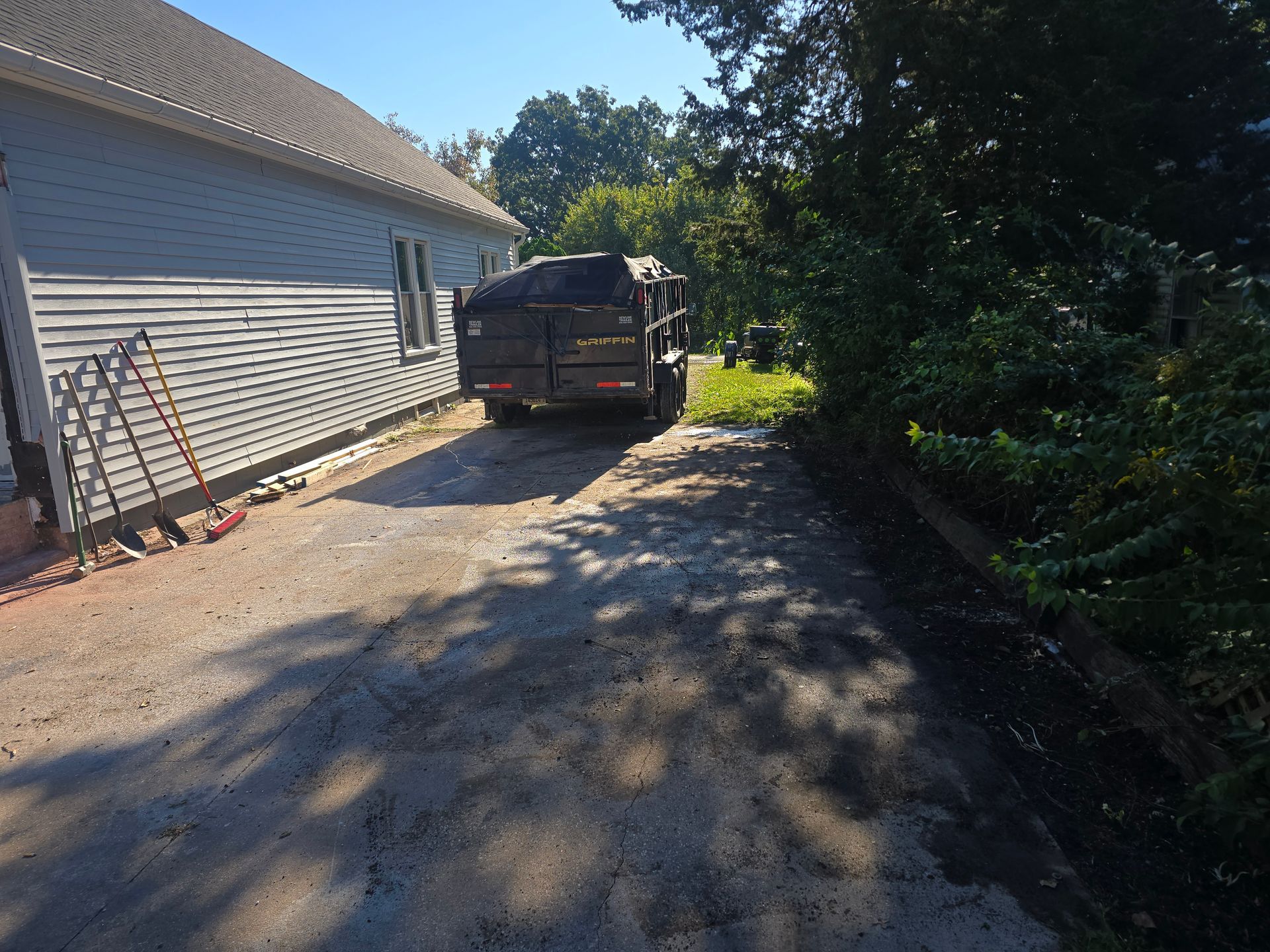 Driveway beside a house with a black trailer parked in sunlight, bordered by trees and bushes
