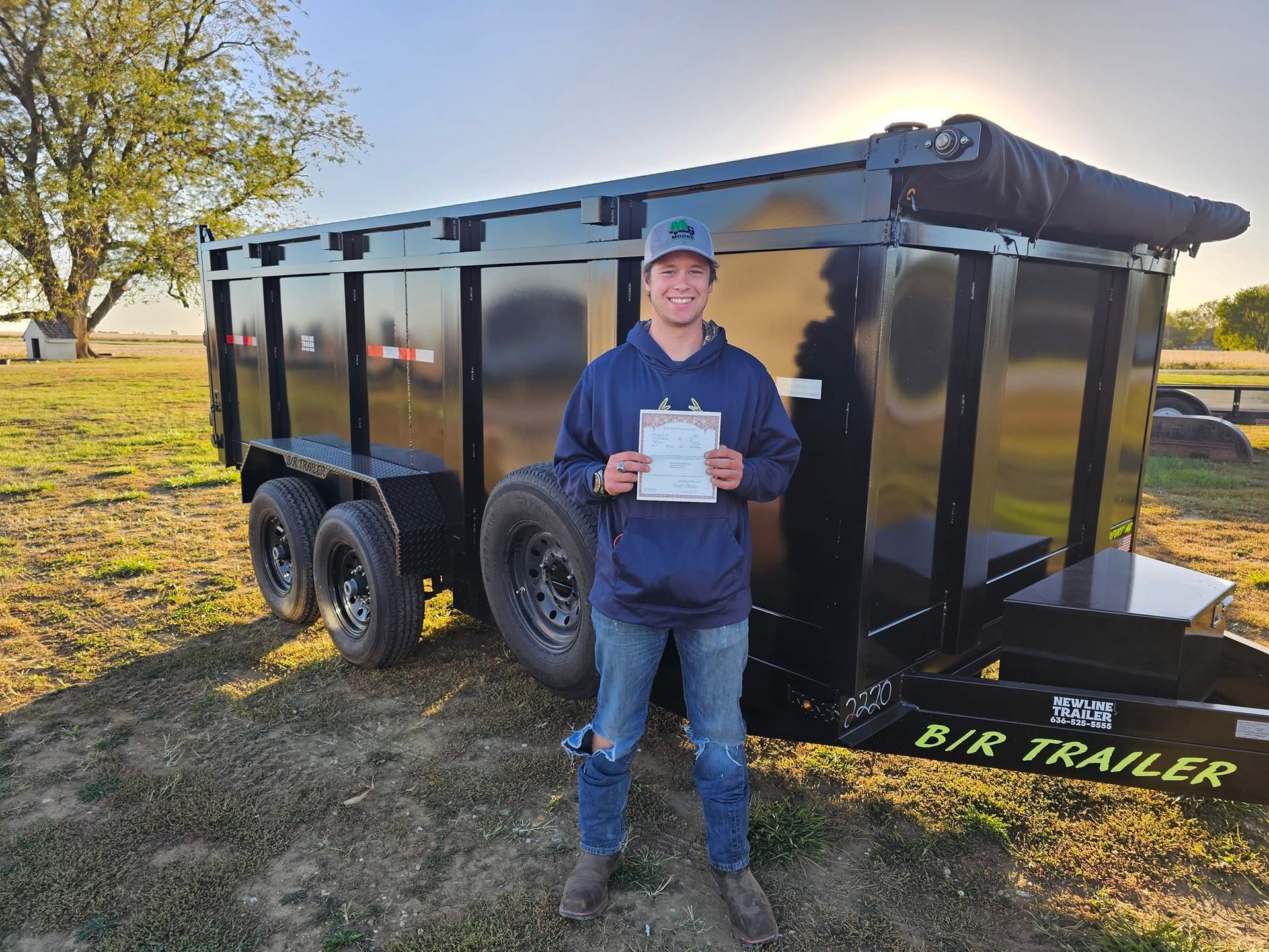 A man is standing in front of a dump trailer holding a clipboard.