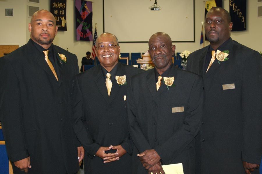 Four men in black suits and gold ties standing together indoors, smiling at the camera.