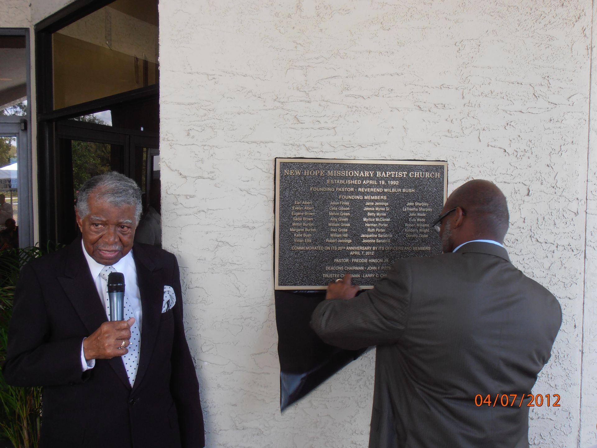 Man speaking at a plaque unveiling beside a stone wall while another man adjusts the plaque.
