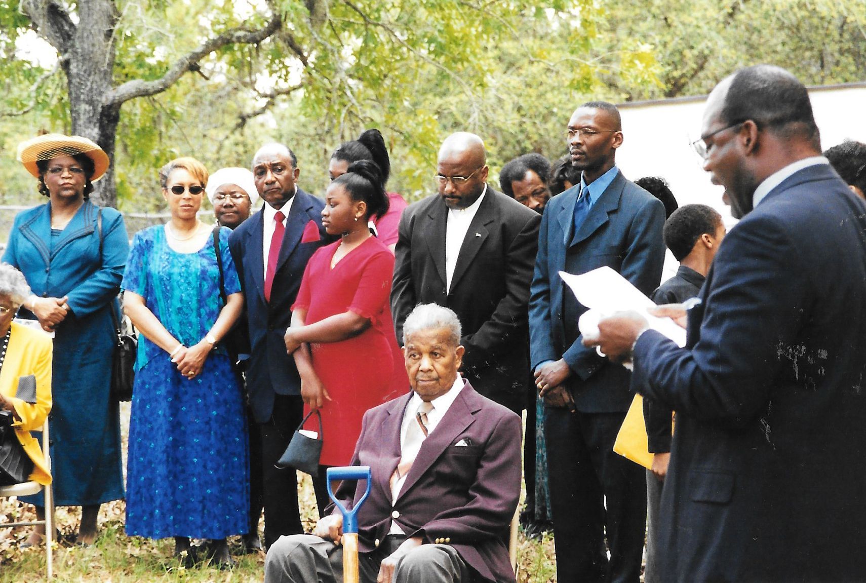 People standing outdoors at a formal gathering, with one man seated in a wheelchair in front