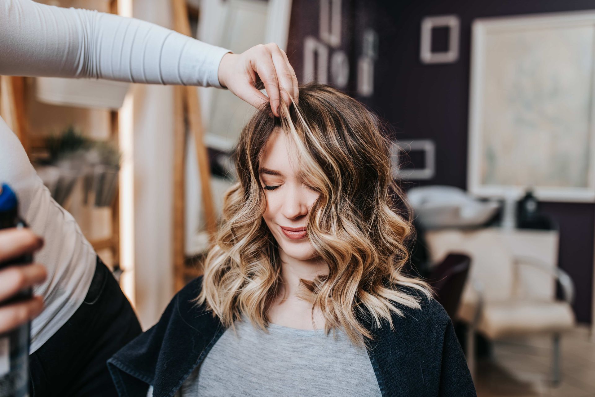 Woman getting hair styled in a salon; hairstylist adjusting hair, wearing gray shirt, salon interior.