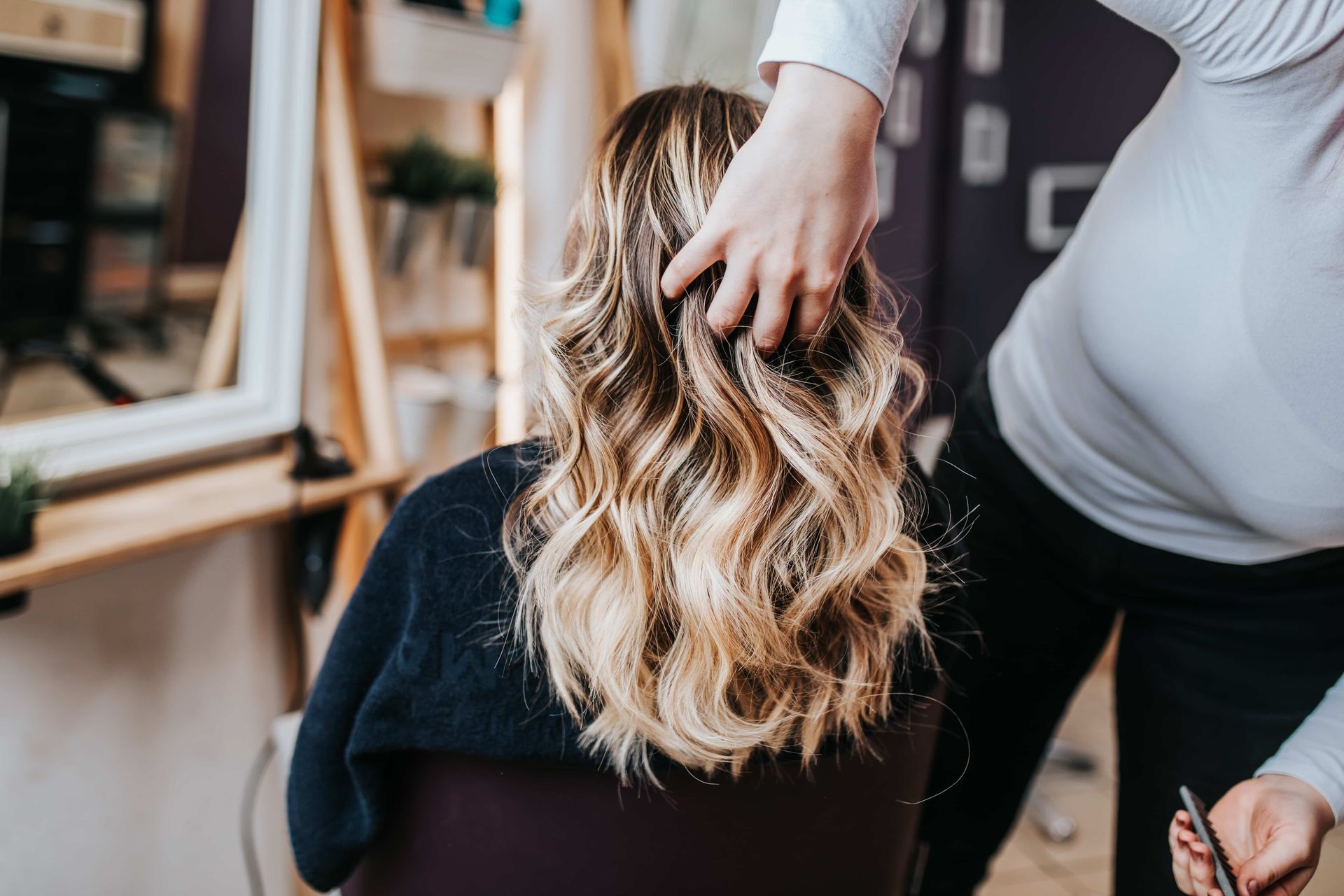 Woman with long wavy blonde hair at a salon, stylist's hands arranging it.