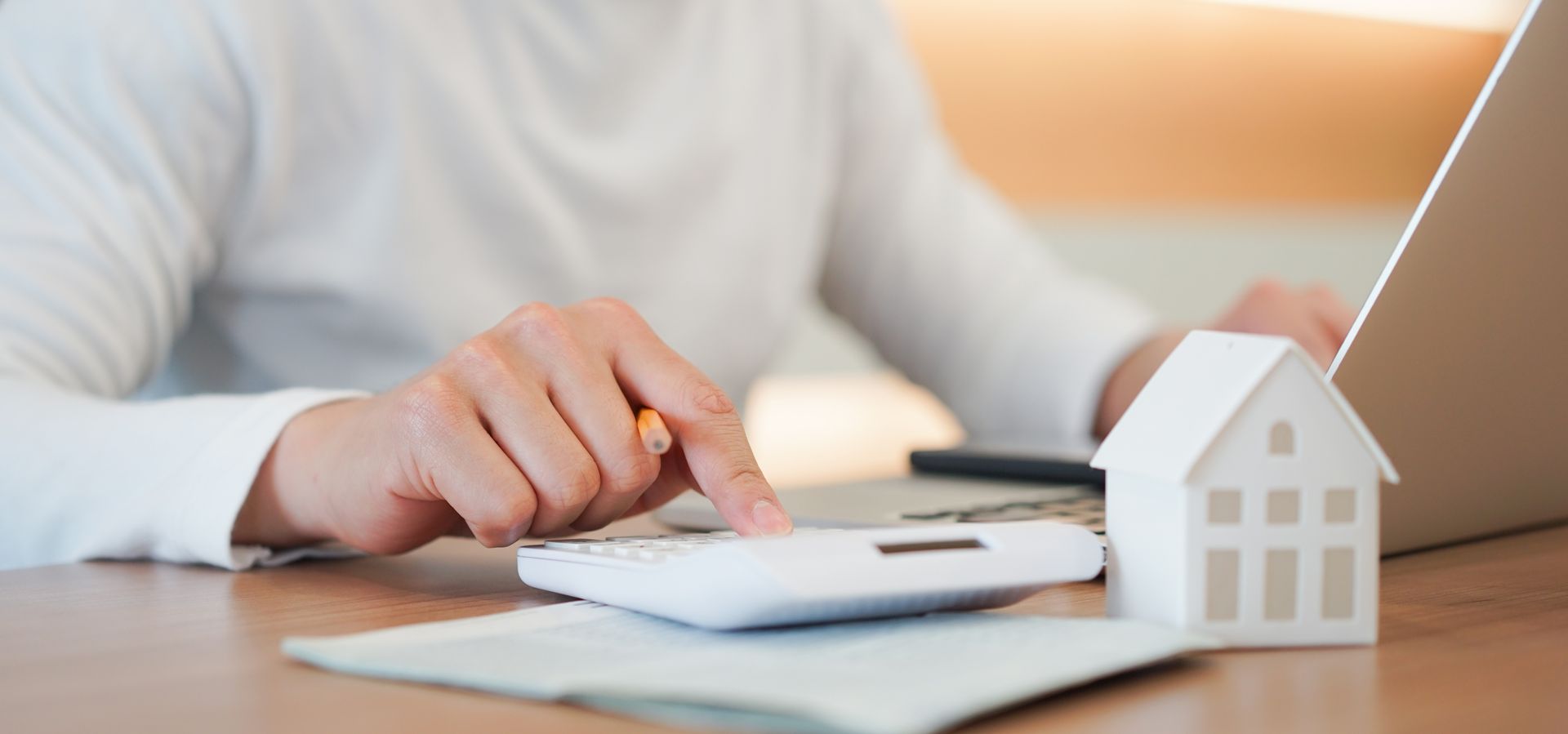 Person using a calculator beside a model house and laptop while reviewing home financing numbers. Person using a calculator beside a model house and laptop while reviewing home financing numbers.