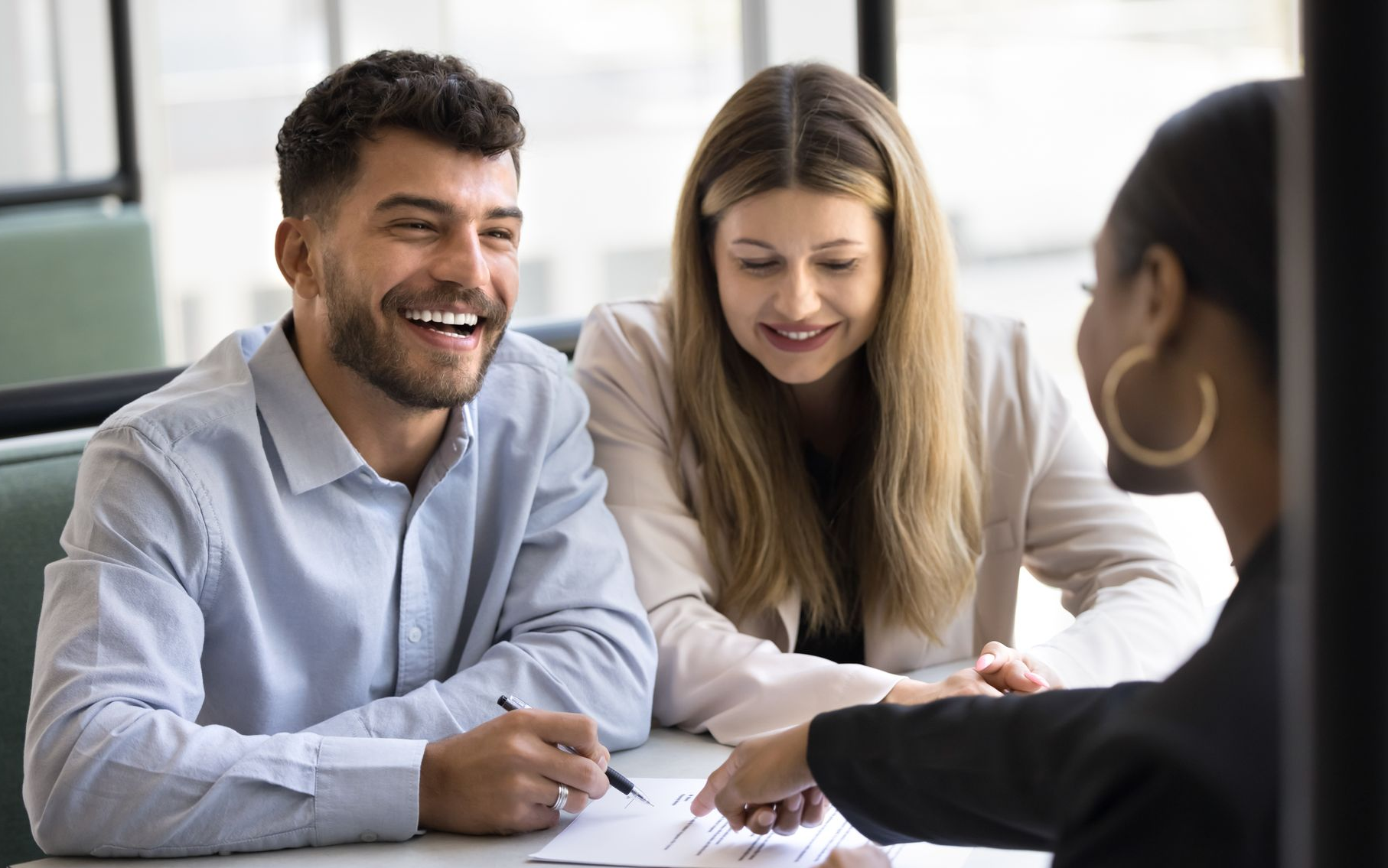 A couple smiling while signing mortgage documents with a loan consultant. Three coworkers smiling and reviewing paperwork at a table in a bright office
