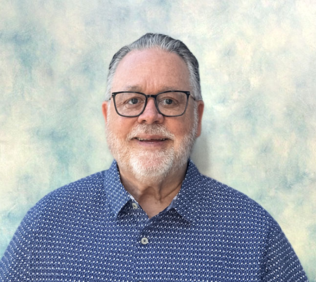 Smiling man with glasses and gray beard in a blue patterned shirt against a soft pastel background