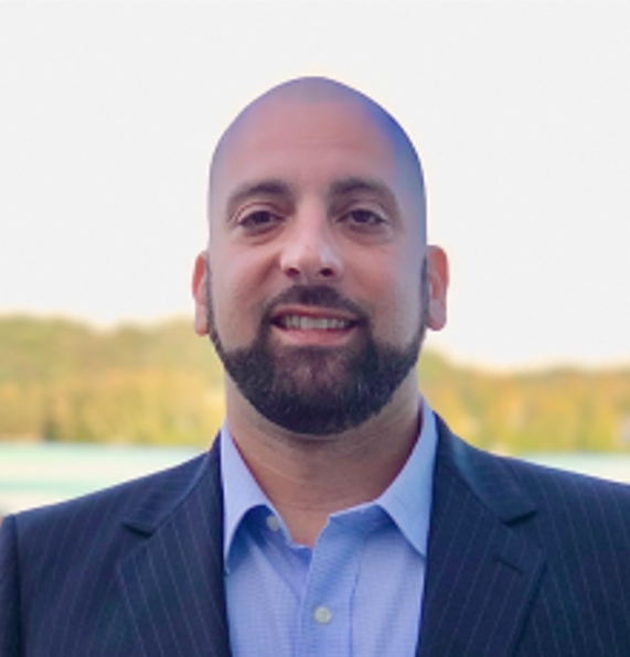 Smiling bald man with a dark beard in a navy suit and blue shirt outdoors, with a blurred landscape behind him