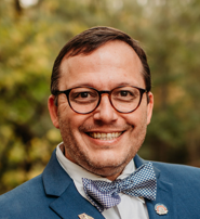 Smiling man in glasses wearing a blue suit and bow tie against a blurred outdoor background