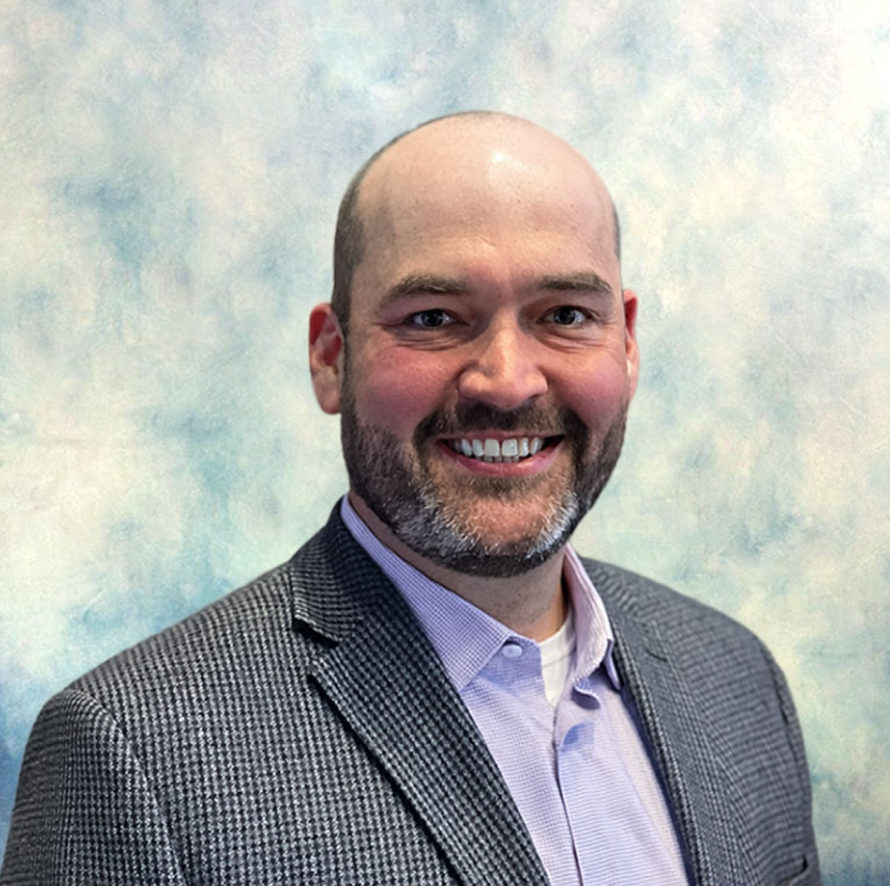 Smiling bald man with a beard in a gray blazer and lavender shirt against a soft blue-gray background