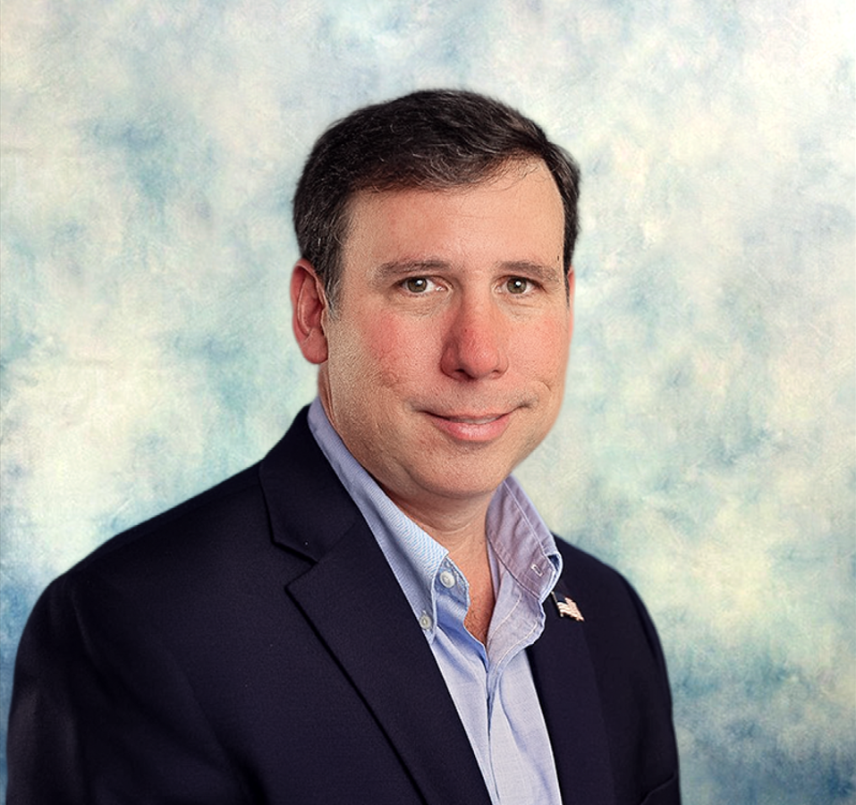 Studio headshot of a man in a navy blazer and light blue shirt against a soft blue background