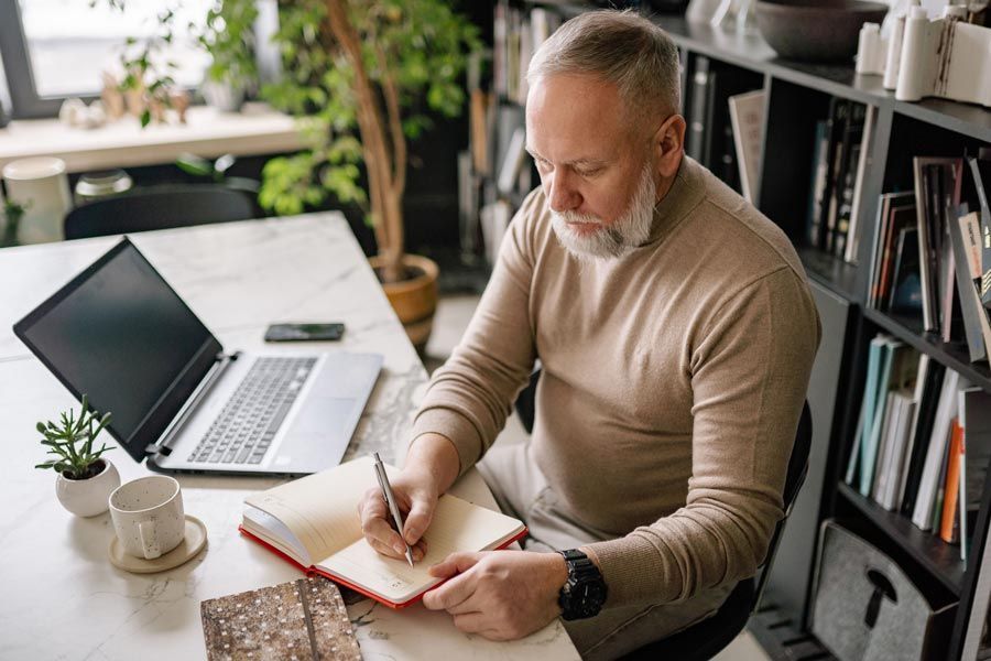 Professional writing in a notebook at a desk with a laptop while planning recruiter outreach