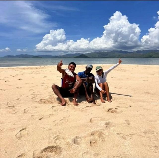 Four people sit on a sandy beach, smiling and waving under a blue sky with large clouds.