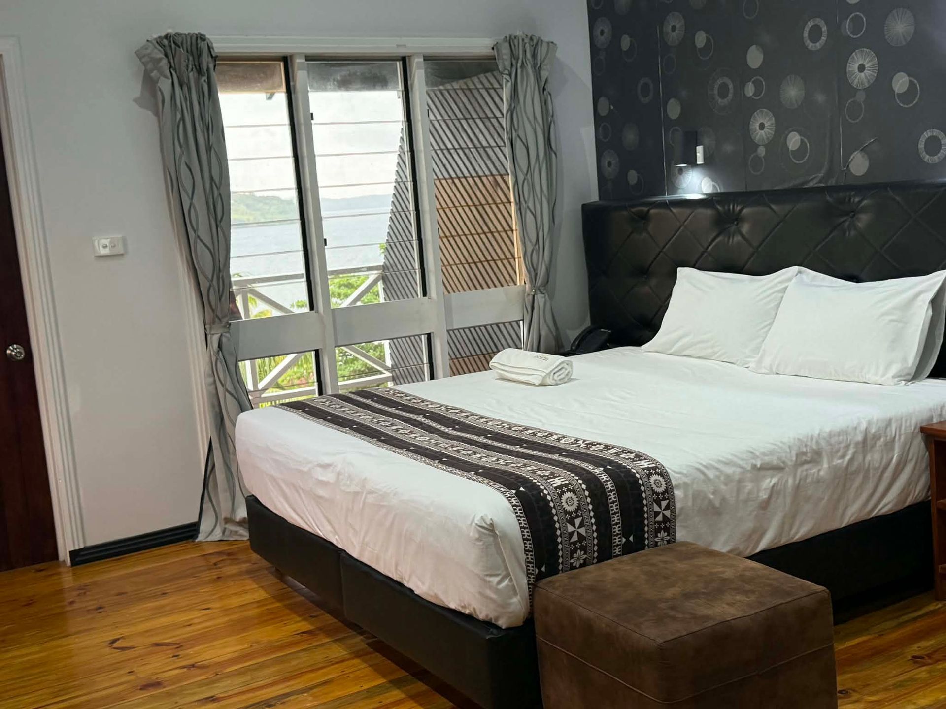 Bedroom with a neatly made bed, dark headboard, brown bench, and patterned curtains by a window