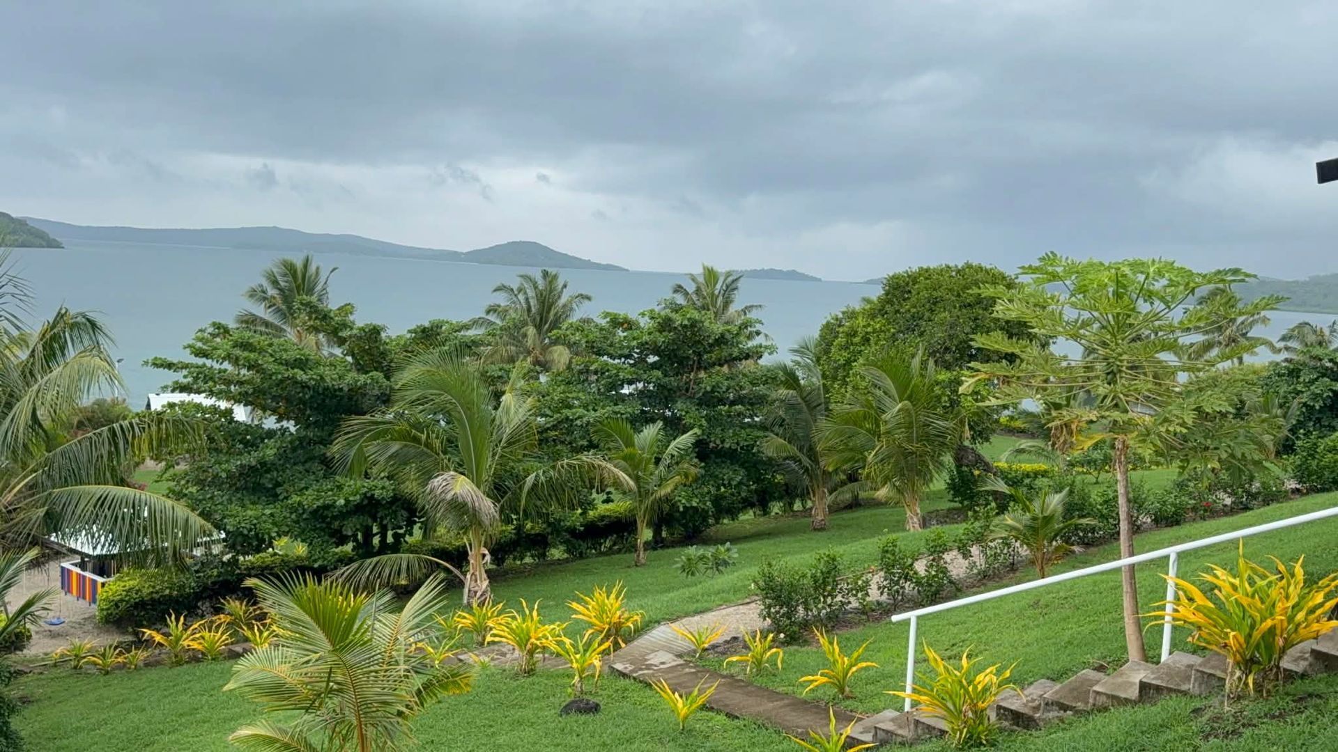 Lush tropical garden with palm trees, flowering shrubs, and mountains under cloudy skies