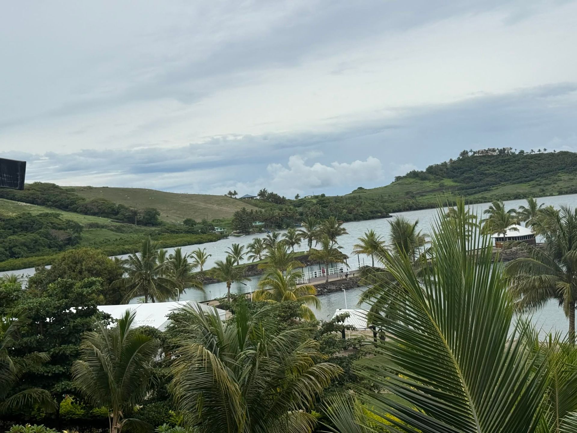 Tropical resort with palm trees, white buildings, and a calm lagoon under a cloudy sky