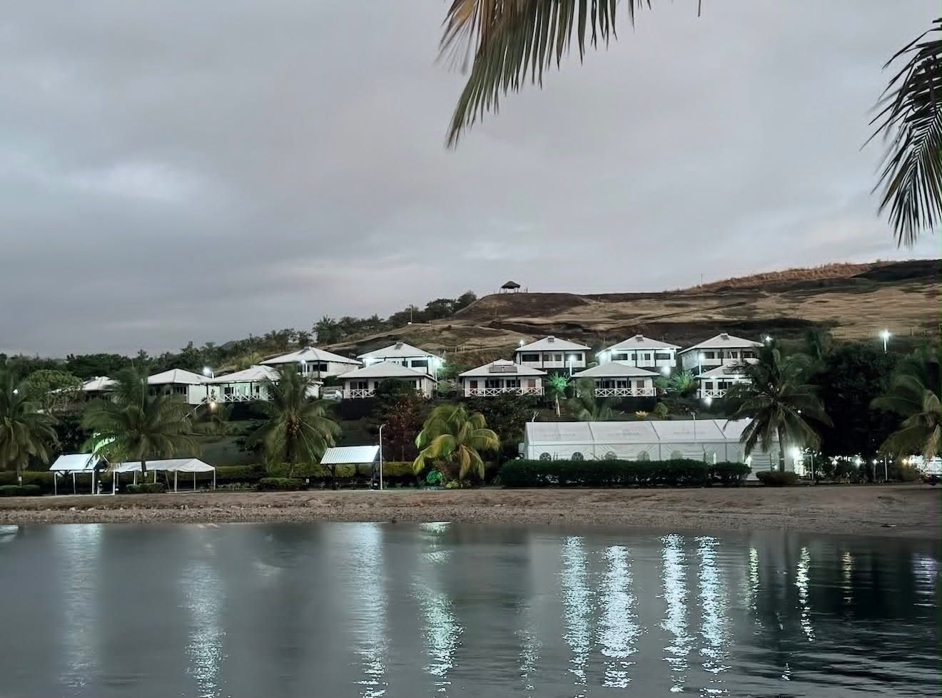 Coastal resort with white buildings, palm trees, and calm water under a cloudy sky