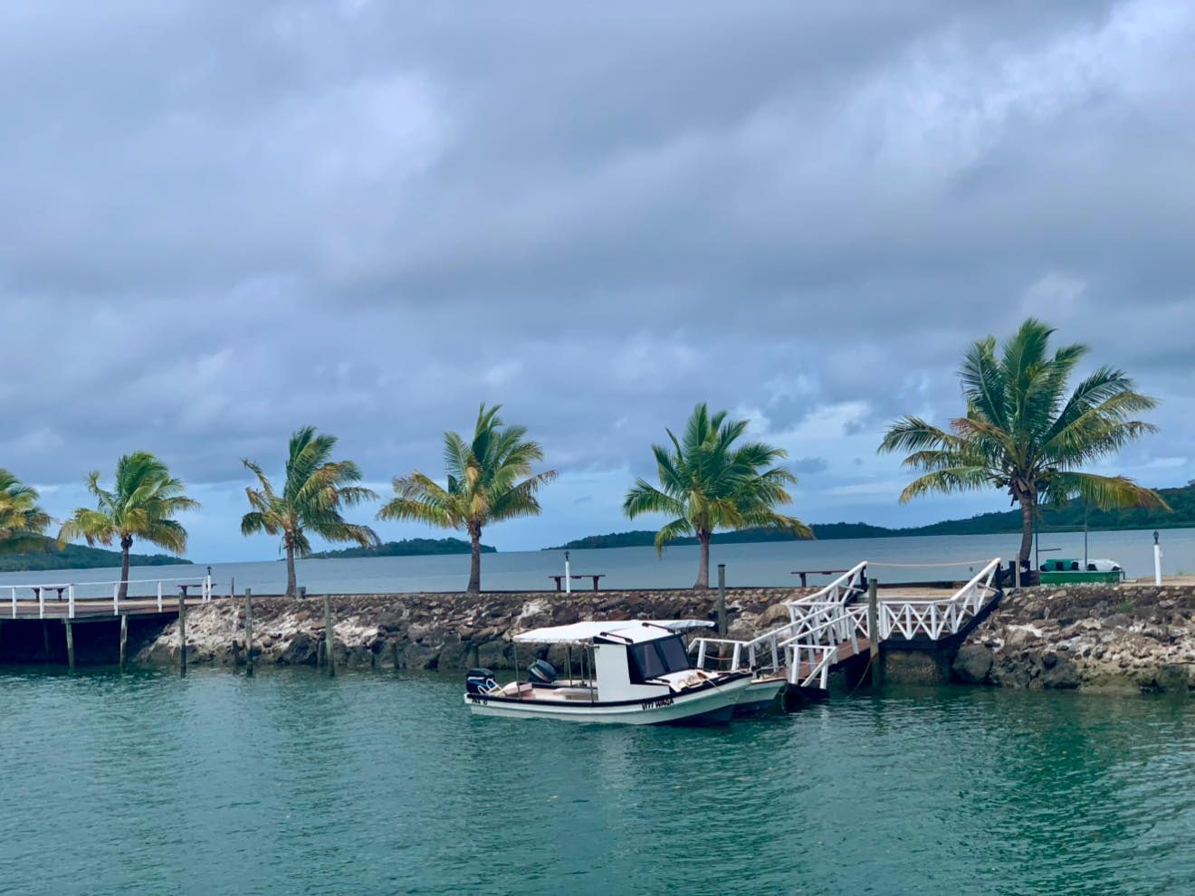 Boat docked by a rocky shore with palm trees, calm turquoise water, and cloudy sky