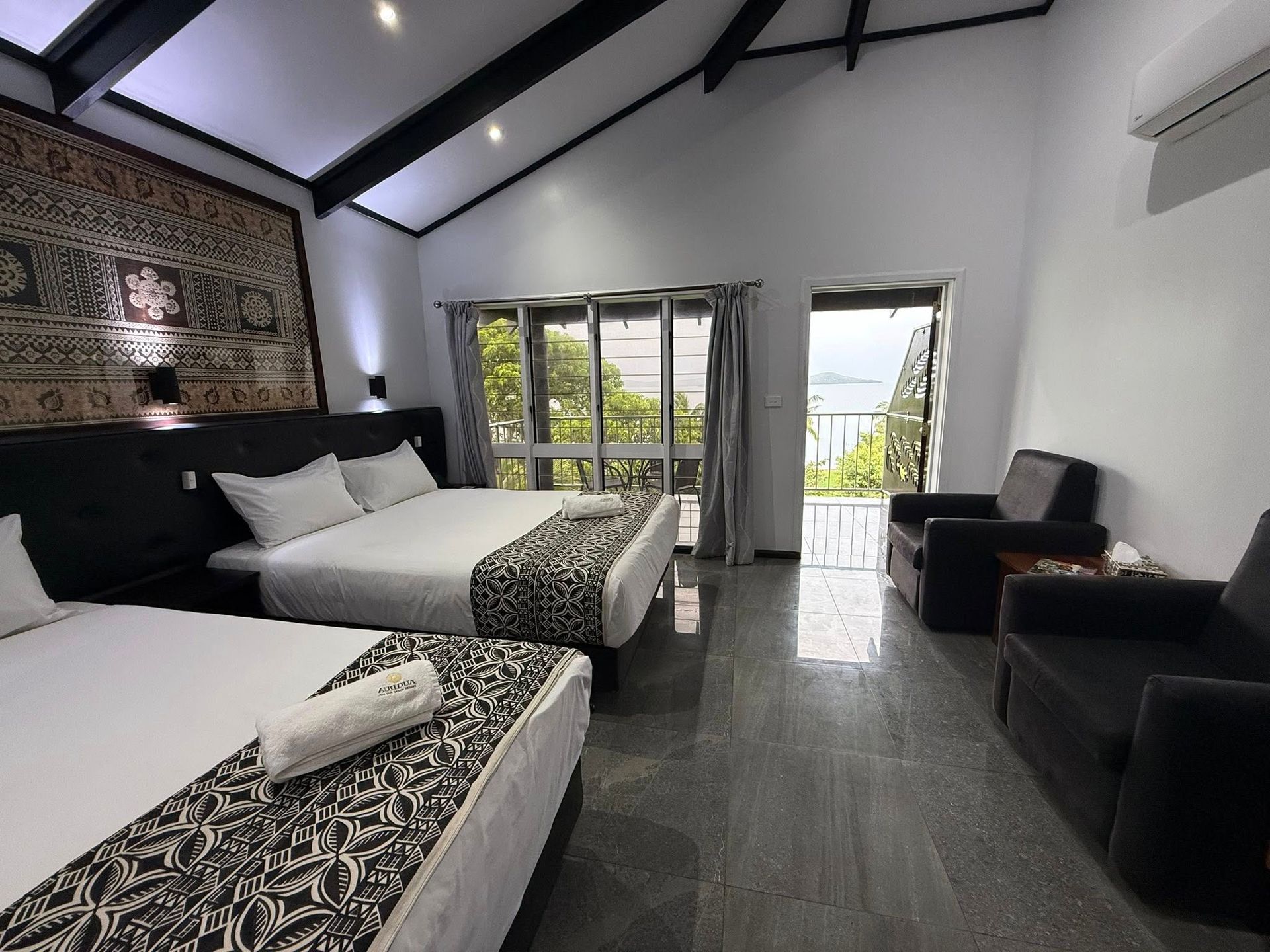 Modern bedroom with two beds, dark chairs, and open doors to a balcony with greenery.