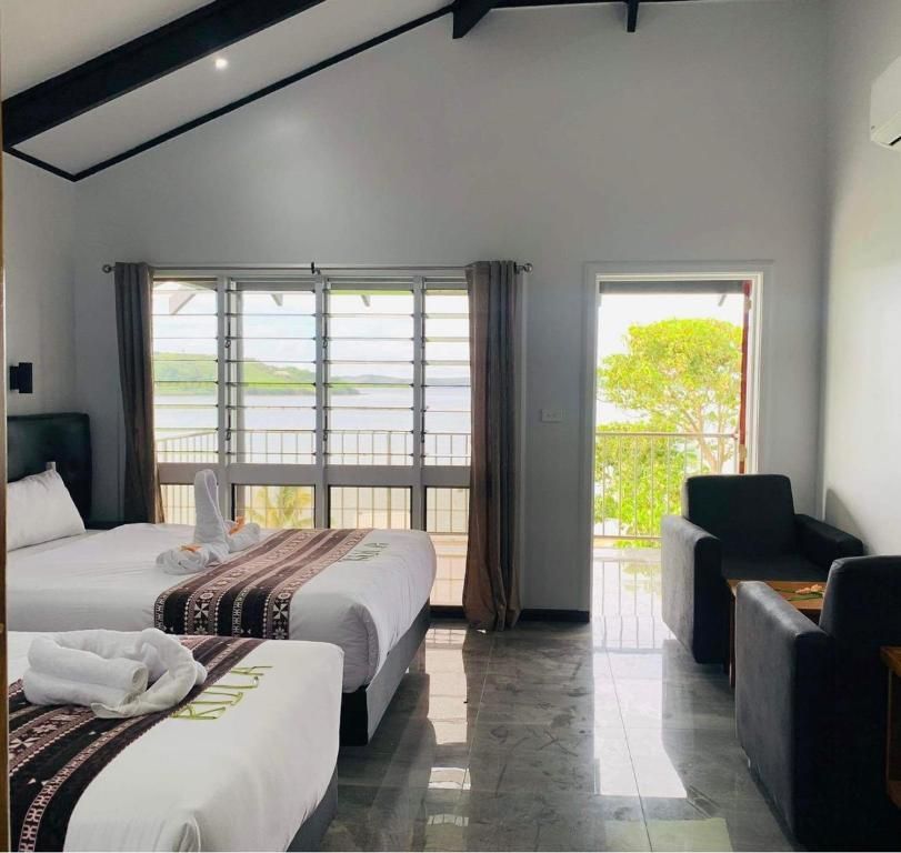 Bright hotel room with two beds, large windows, dark curtains, and a seating area overlooking water