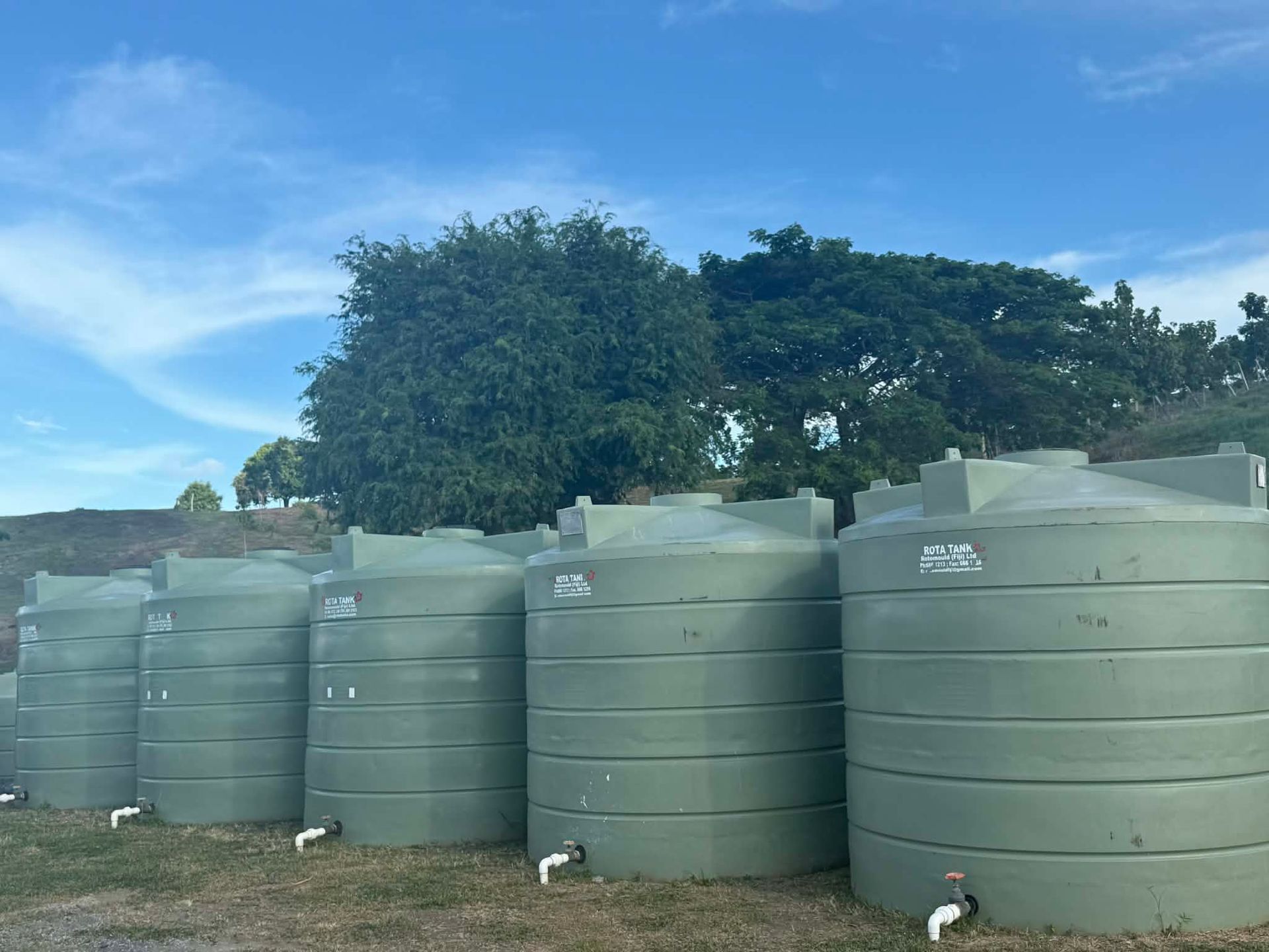 Row of large green water tanks outdoors under a blue sky with trees behind them