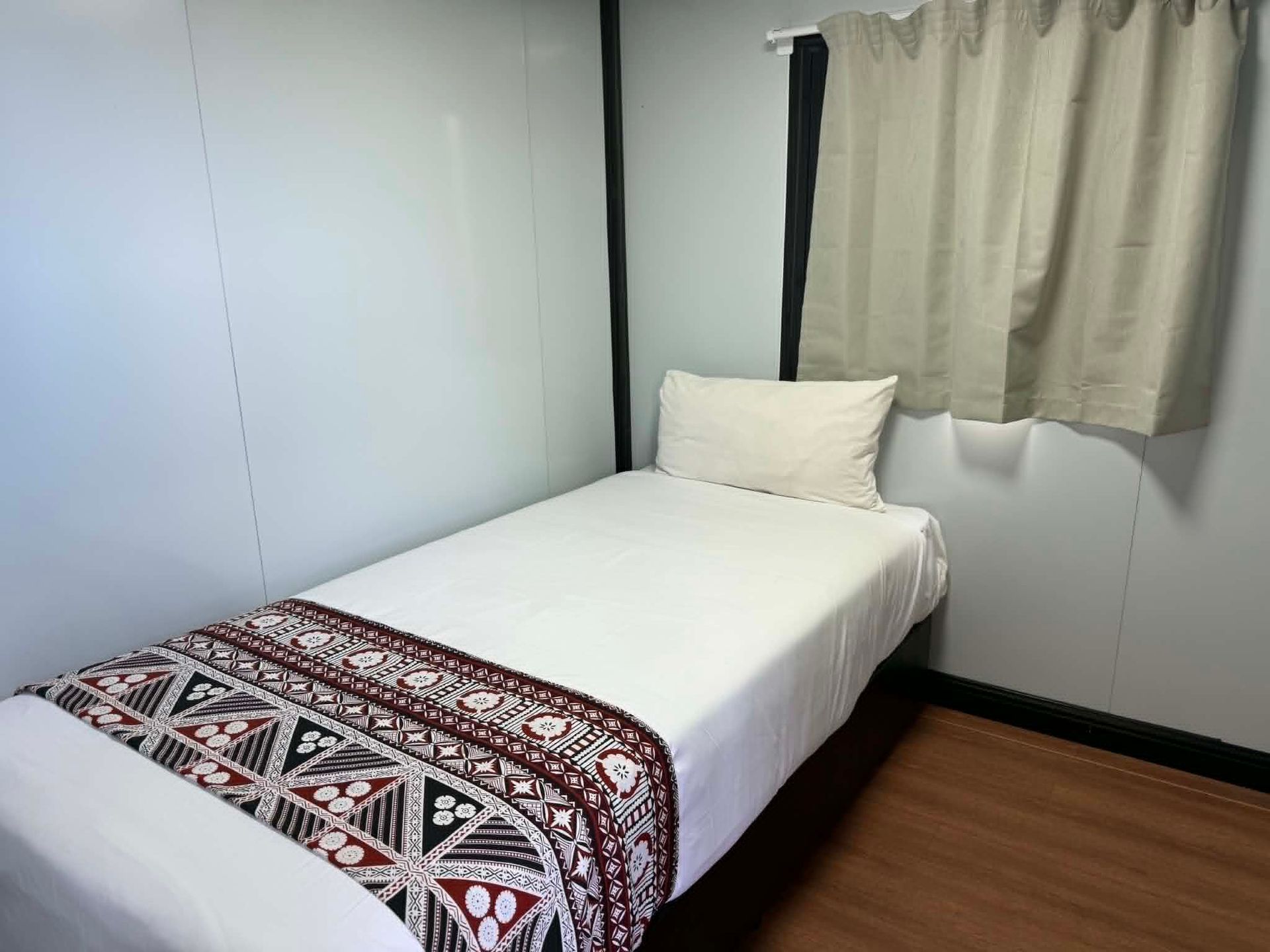 Small bedroom with a single bed, patterned blanket, white walls, and a curtained window.