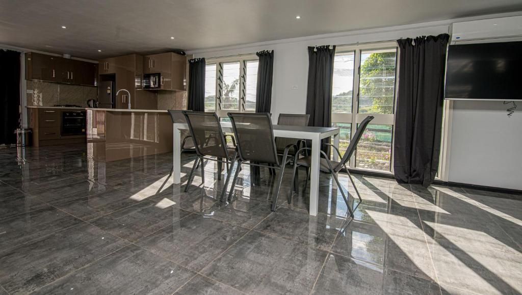 Modern open-plan kitchen and dining area with dark flooring, glass table, chairs, and large windows with curtains