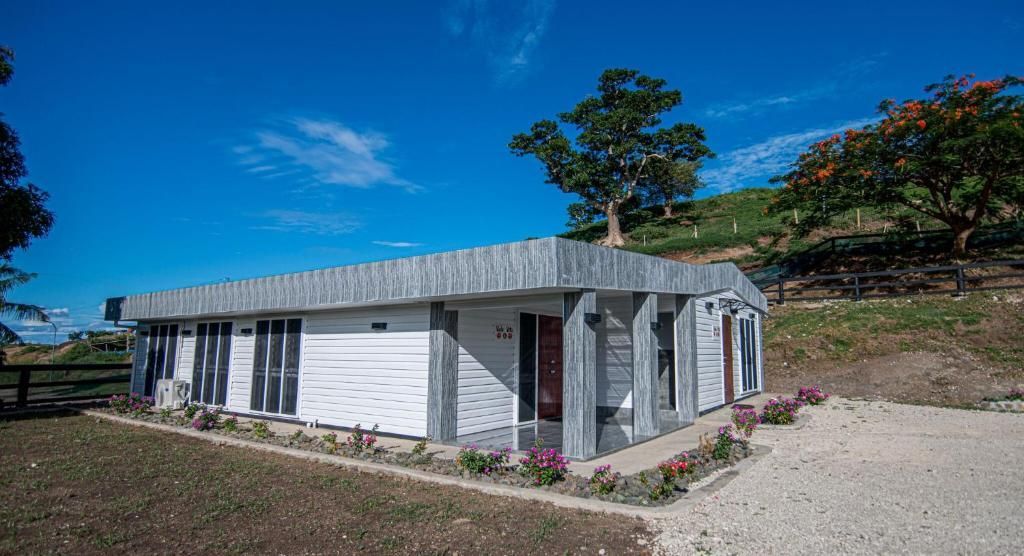 Small white house with a gray roof on a gravel lot under a bright blue sky, beside a hillside tree