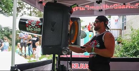 DJ at a booth under an umbrella, speaking into a microphone beside a large speaker outdoors.