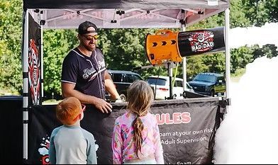 Man under a canopy booth talks to two children, with posters and a black banner behind him.