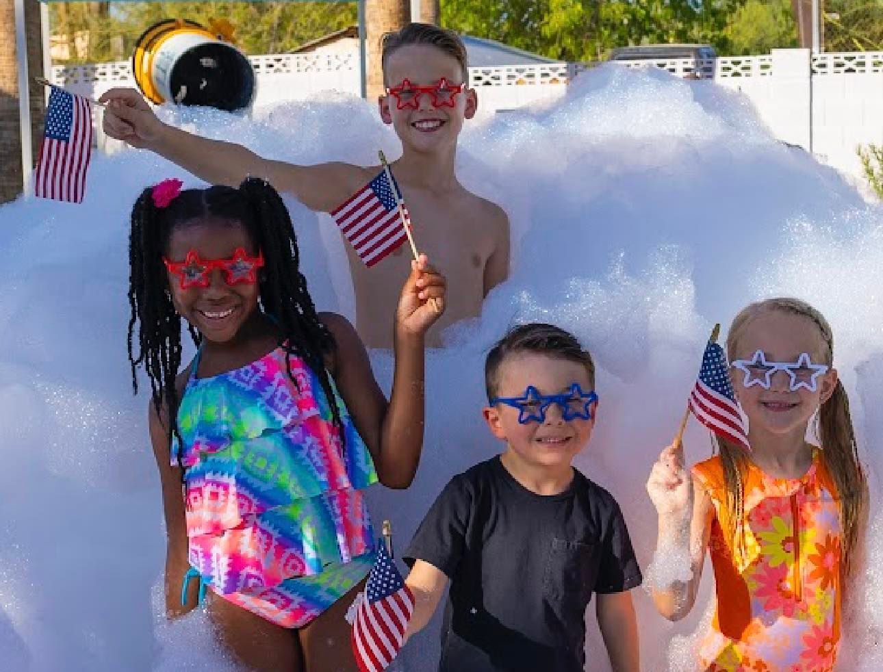 Four children in sunglasses hold small American flags outdoors by a snowy mound.
