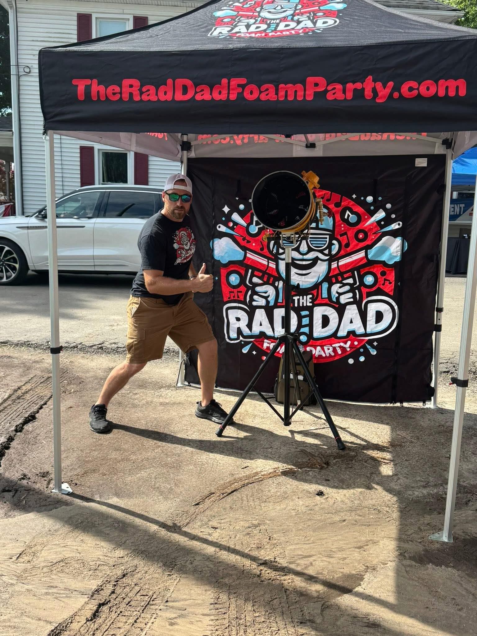 Man posing beside The Raw Dog Foam Party canopy and banner outdoors on gravel.