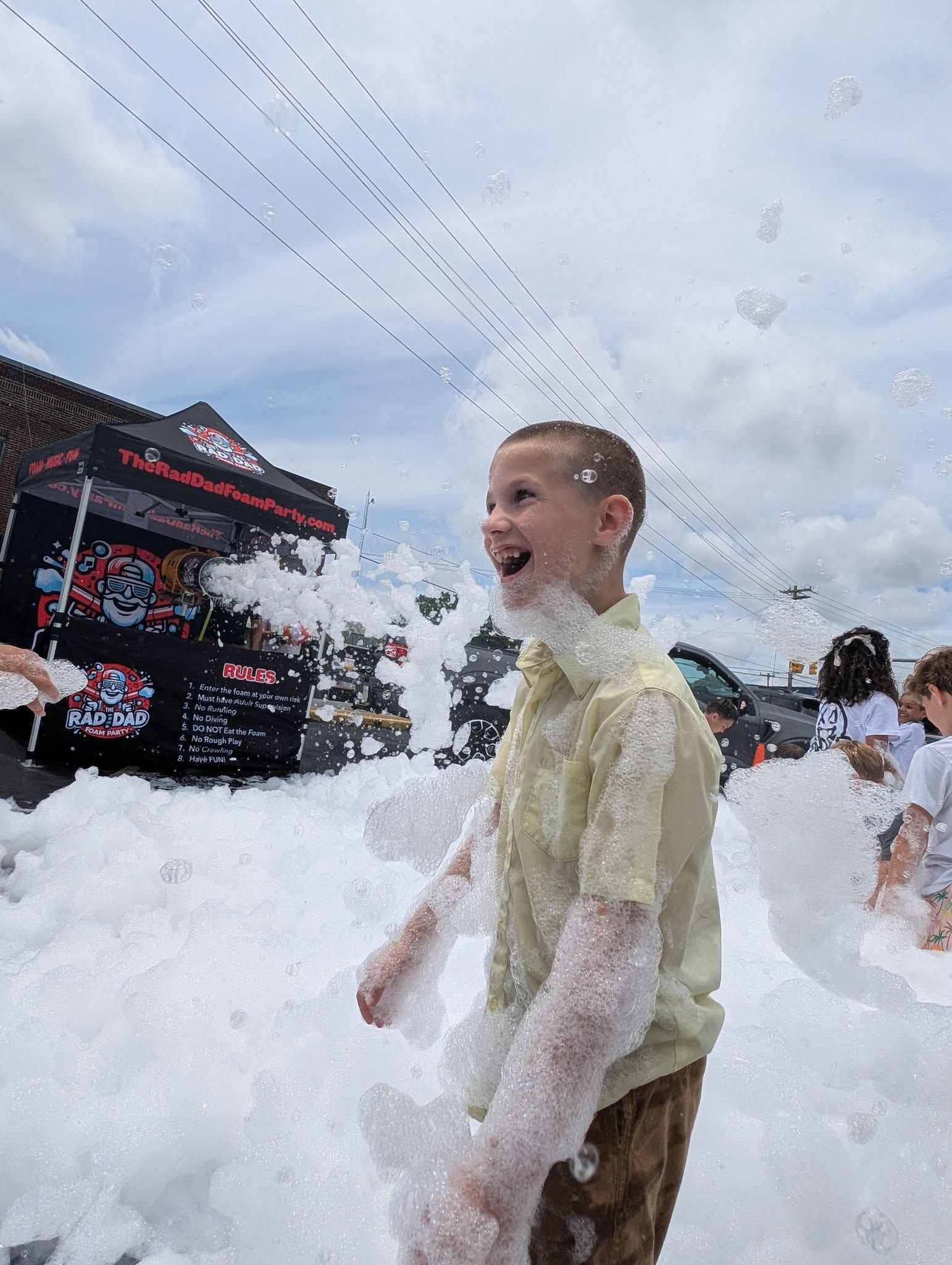Person smiling amid foam at an outdoor event, with a red-and-black stage in the background
