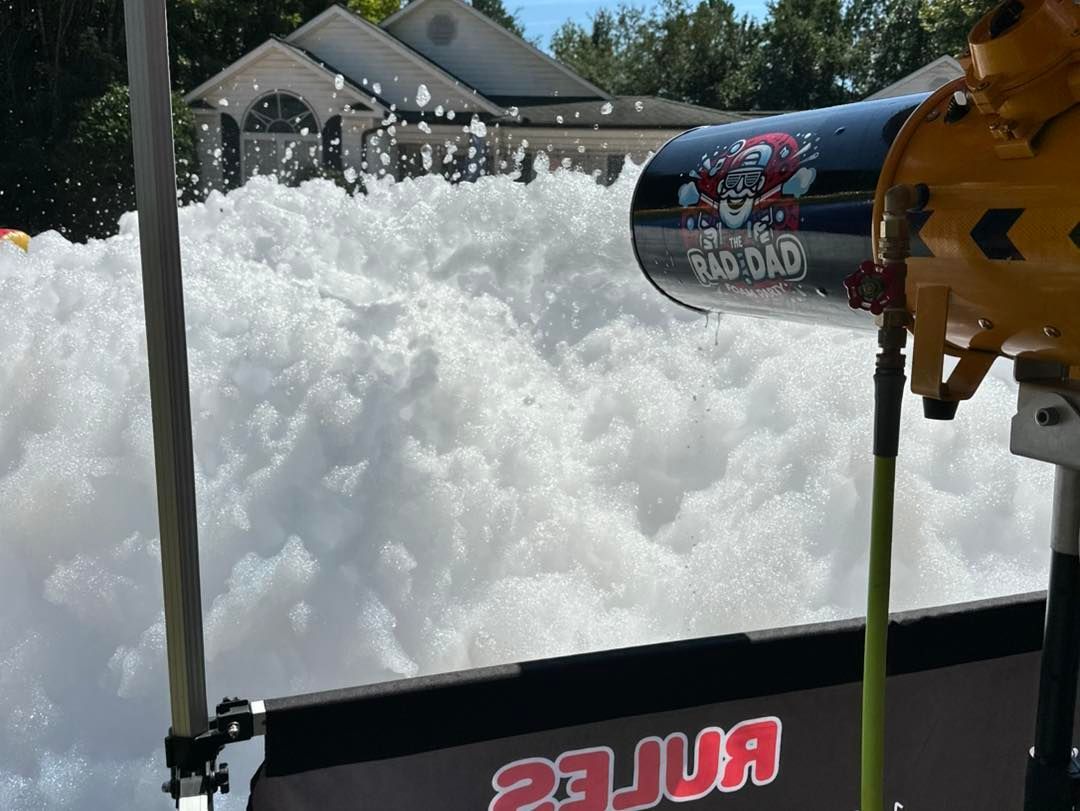 Snow sprayed across a suburban yard from a hose or blower with a yellow nozzle.
