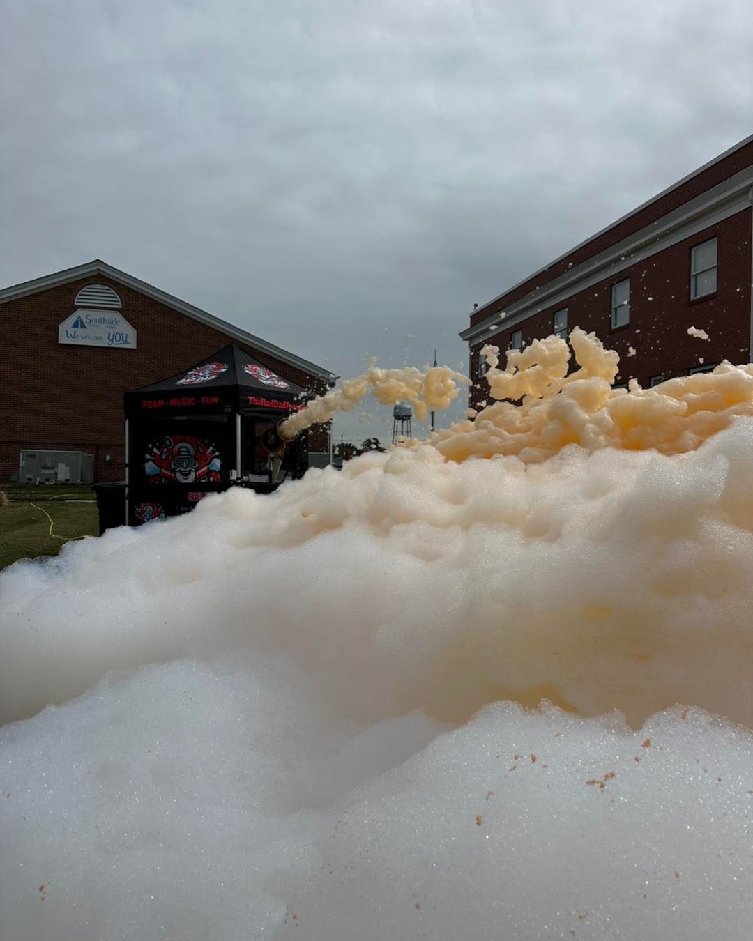 Foamy white floodwater piled high beside a brick building and parking lot under a gray sky