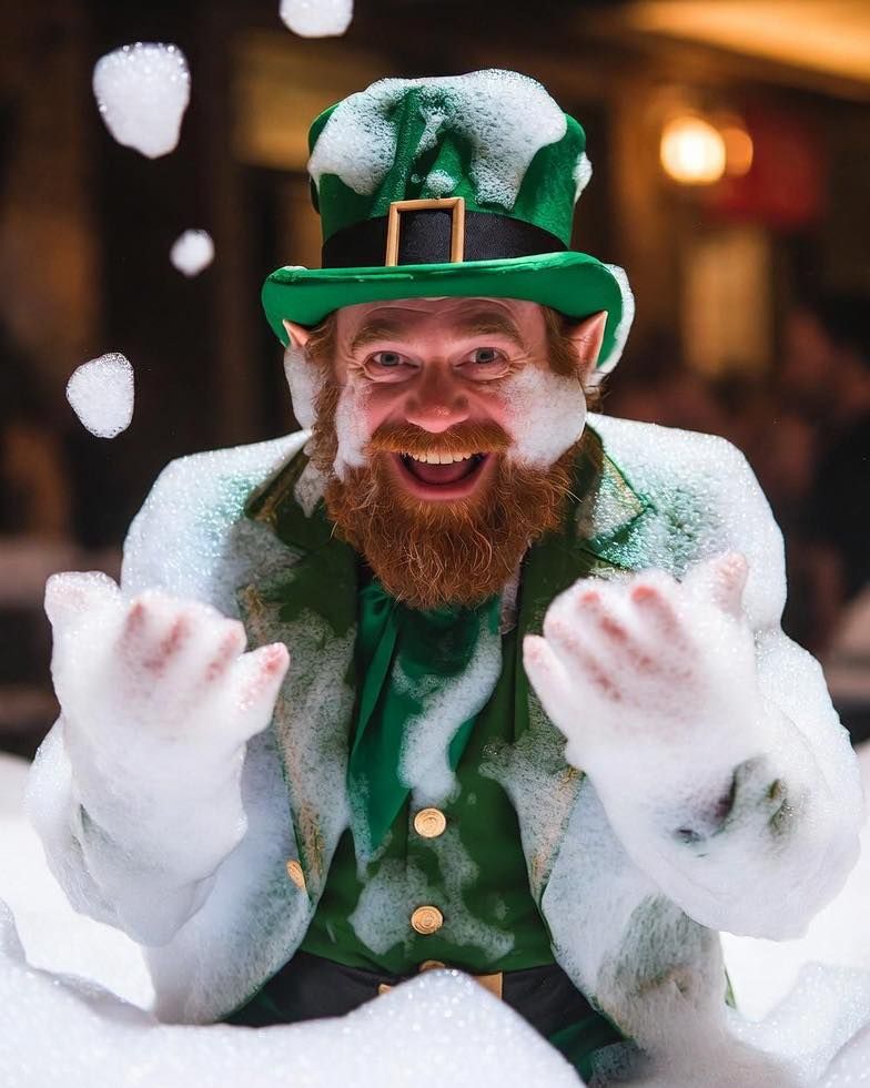 Smiling bearded man in a green leprechaun outfit and hat, holding up snow-covered hands.