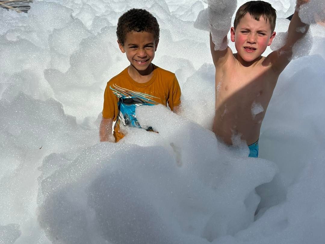 Two children playing in a pile of foam, one in an orange shirt and one shirtless, smiling outdoors