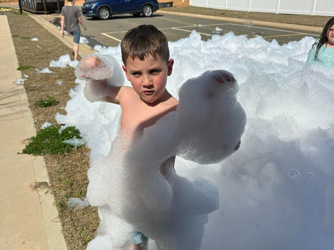 Child playing in a large pile of white foam outdoors, reaching toward the camera.