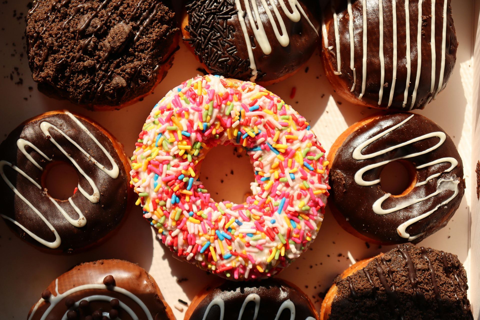 Assorted chocolate and sprinkled donuts, with a pink sprinkled donut centered on a warm brown background