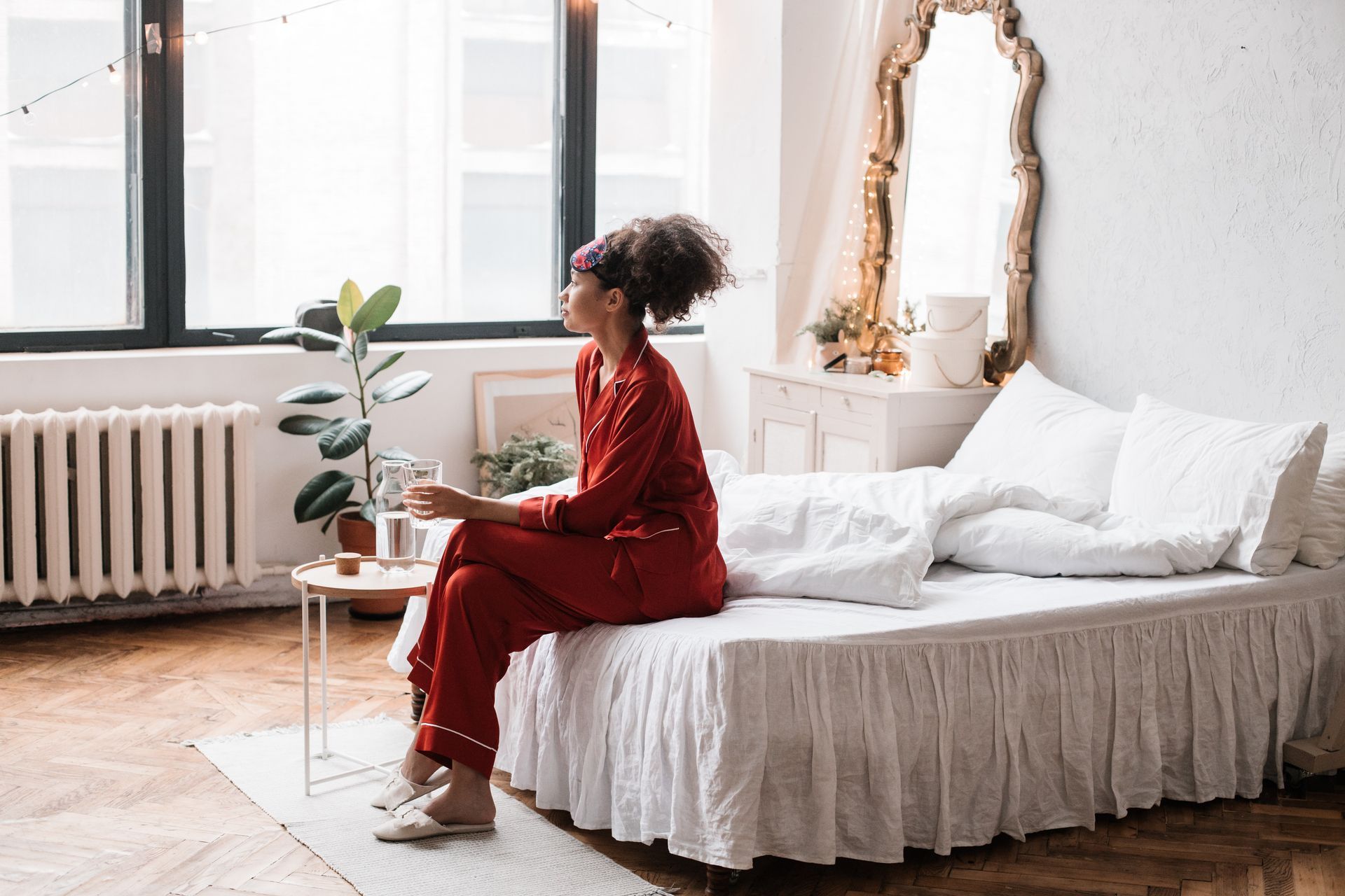 Woman in red pajamas sits on bed near a window, looking outside. Bright room, with mirror and plant.