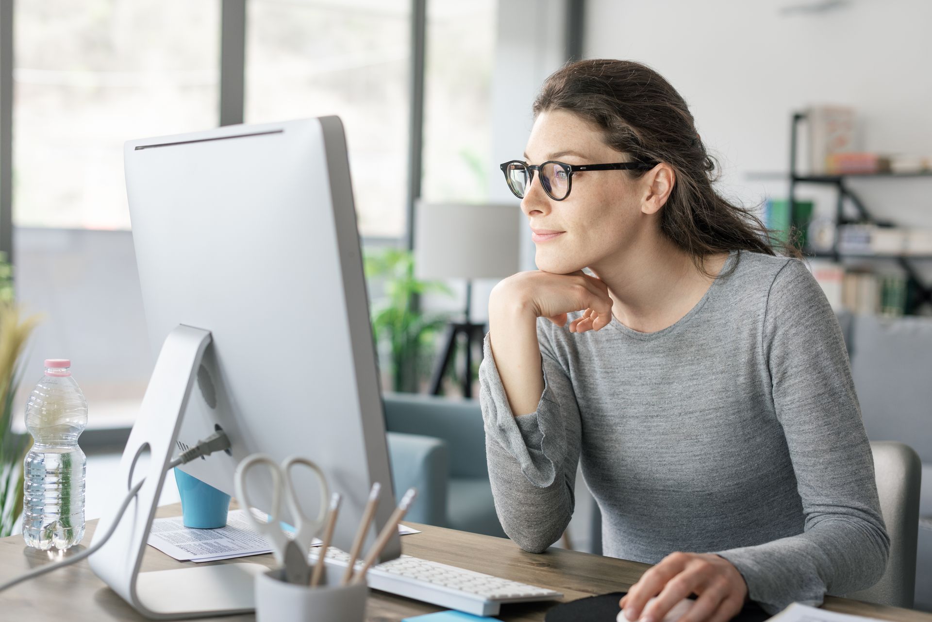 Woman with glasses working on a computer at a desk, looking thoughtfully.