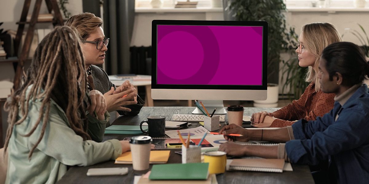 Four people at a table, looking at a purple computer screen, discussing ideas. Inside a modern office setting.