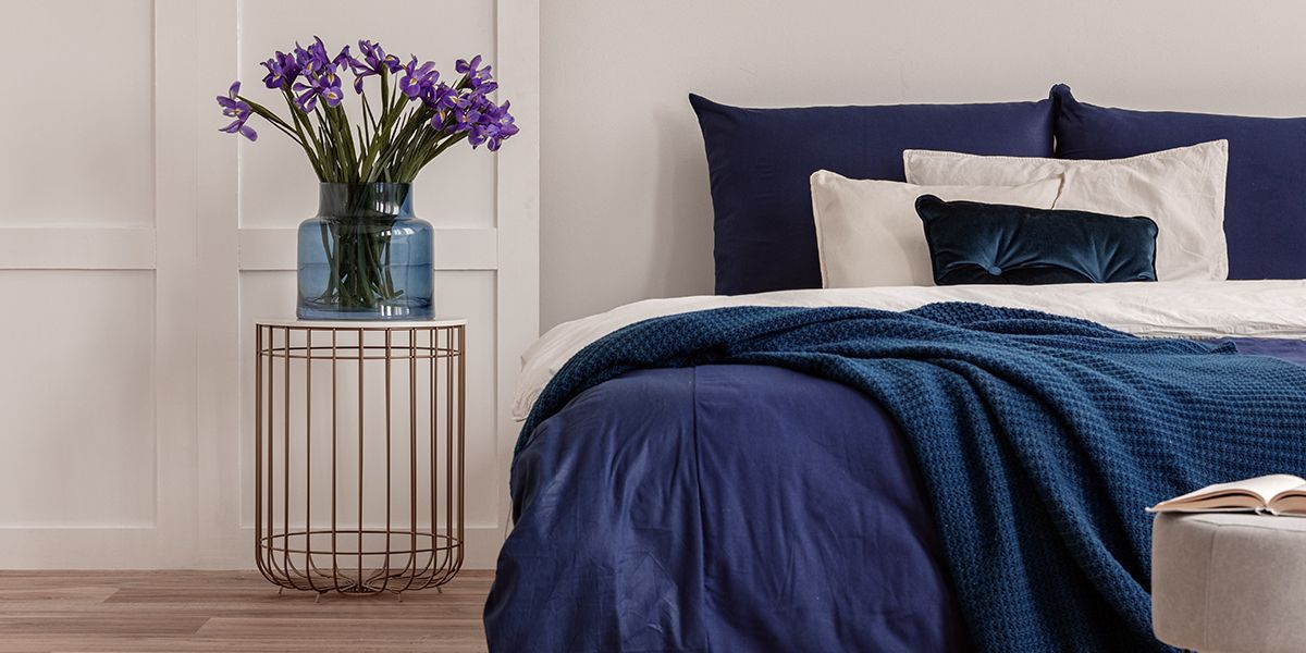 Bedroom with blue bedding, a vase of purple flowers, and a side table.