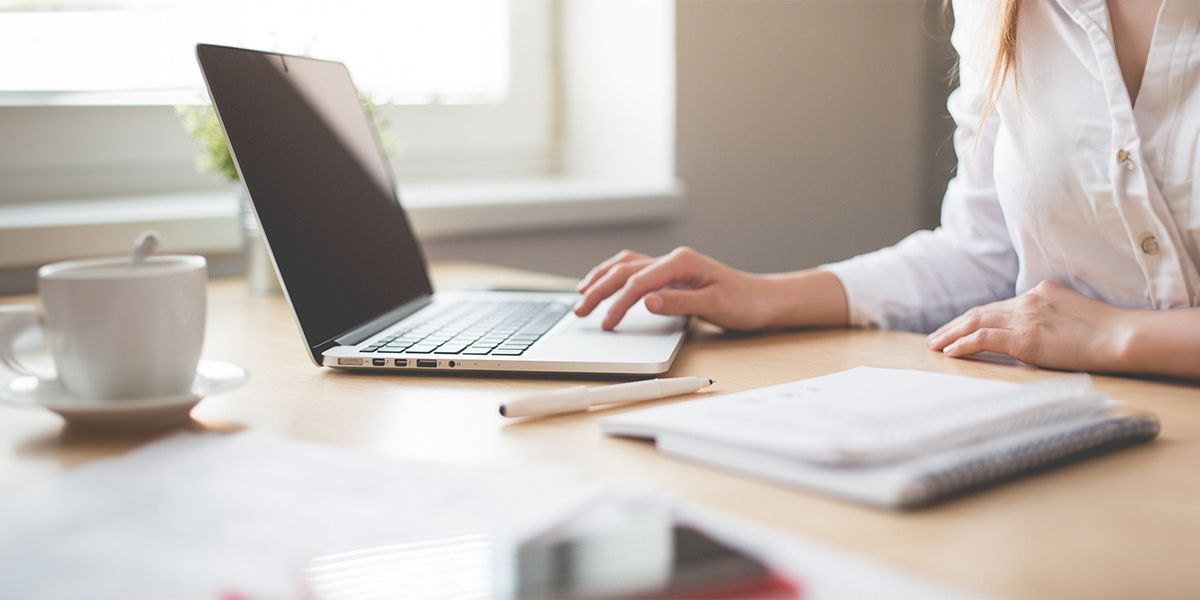 A person working on a laptop at a desk with a cup of coffee, pen, and notepad.