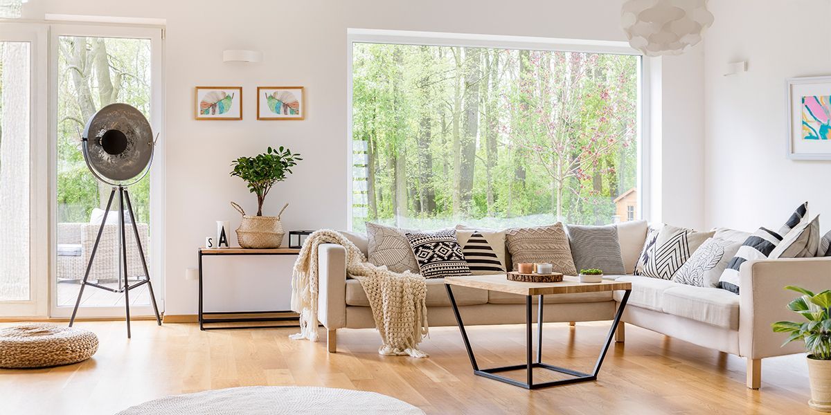Bright, airy living room with a large window overlooking a forest. Beige sofa with decorative pillows, wooden floors.