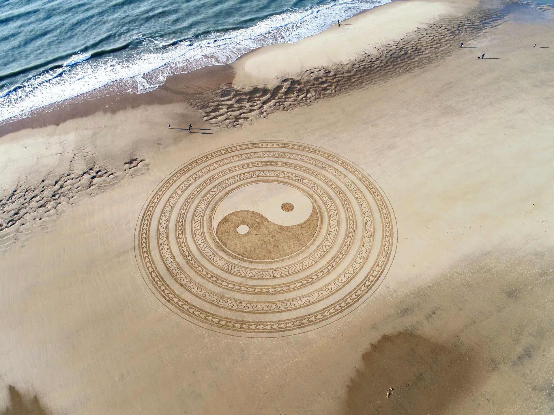 Yin and Yang symbol sand art on a beach, with ocean waves in the background.