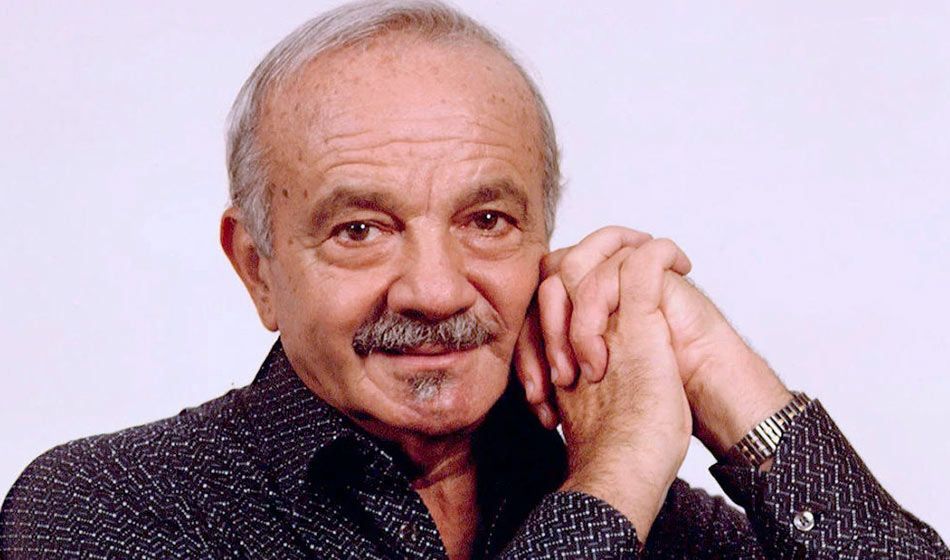 Hombre con cabello canoso, bigote y perilla, con las manos entrelazadas cerca de la cara, mirando a la cámara.