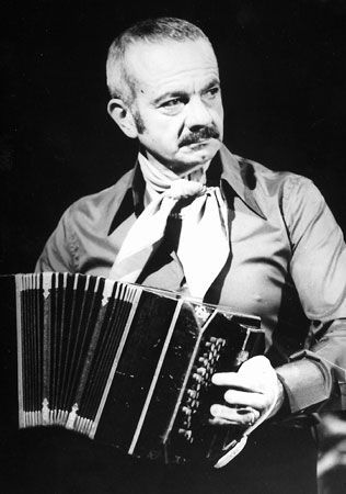 Hombre con bigote tocando un bandoneón, vistiendo una bufanda y una camisa abotonada.